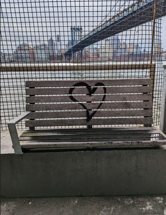 A wooden bench, resting on concrete, with water and city views behind. A heart drawn on the back of the bench.