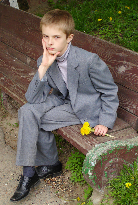 A young boy in a grey suit, lavender coloured shirt, holding a dandellion, sitting on a bench