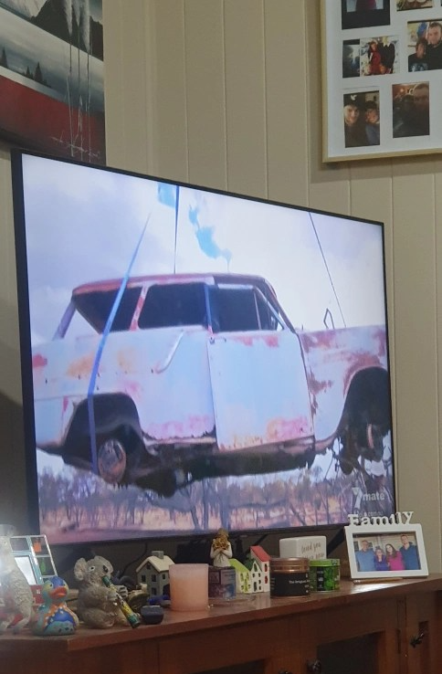 A big screen TV with an image of a rusty old car, being raised with straps.