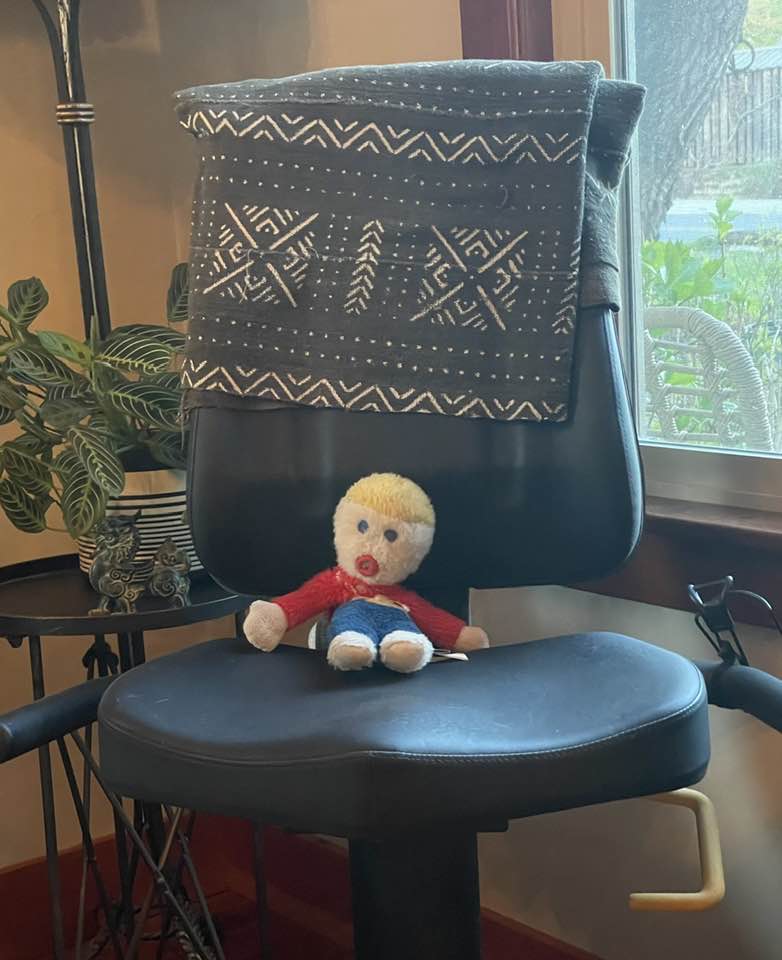 Small stuffed toy, bald yellow head, red jacket and blue pants, sits on an office chair. There is a blanket on the back of the chair. And an indoor plant and view of the garden.